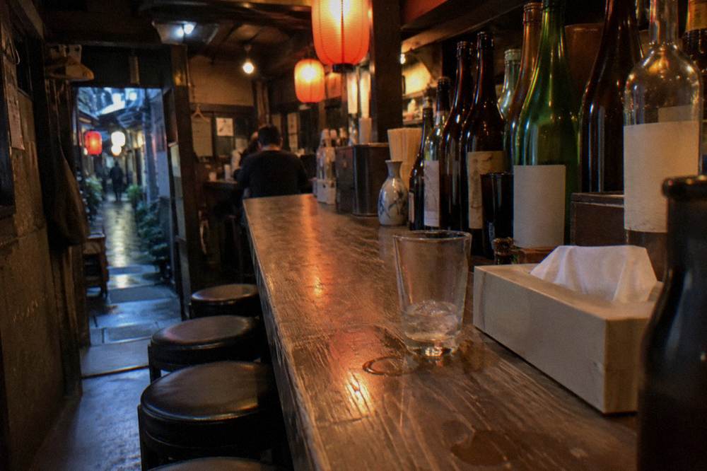 Small lantern-lit izakaya interior with bottles lined along a wooden counter