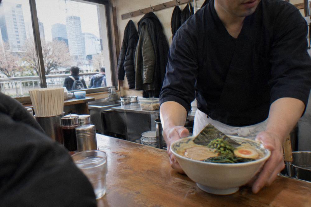 A busy Tokyo ramen counter with a chef placing a bowl on the counter