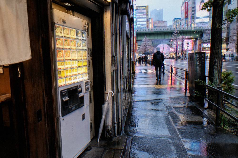 Close view of a ticket machine menu showing ramen bowls and prices beside a shop entrance