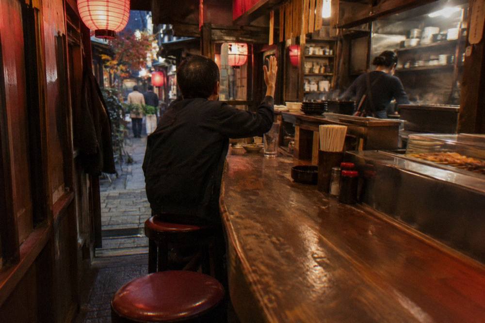 A diner raising one hand to signal a server across a warm lantern-lit izakaya counter