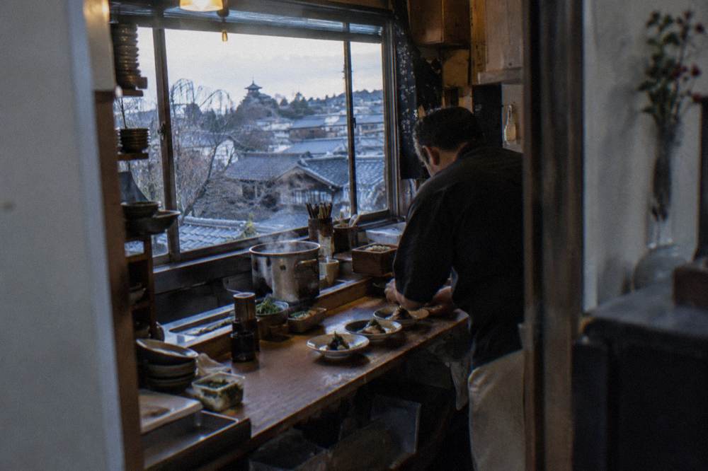 A chef plating small dishes across a wooden restaurant counter in the evening