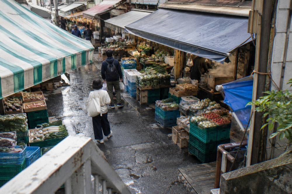 Morning light over narrow market lanes with awnings, stacked crates and shoppers walking between stalls