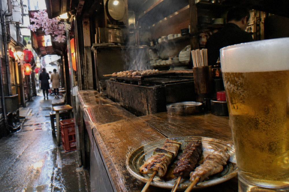 Small grilled skewers and a glass of beer on a worn wooden bar counter