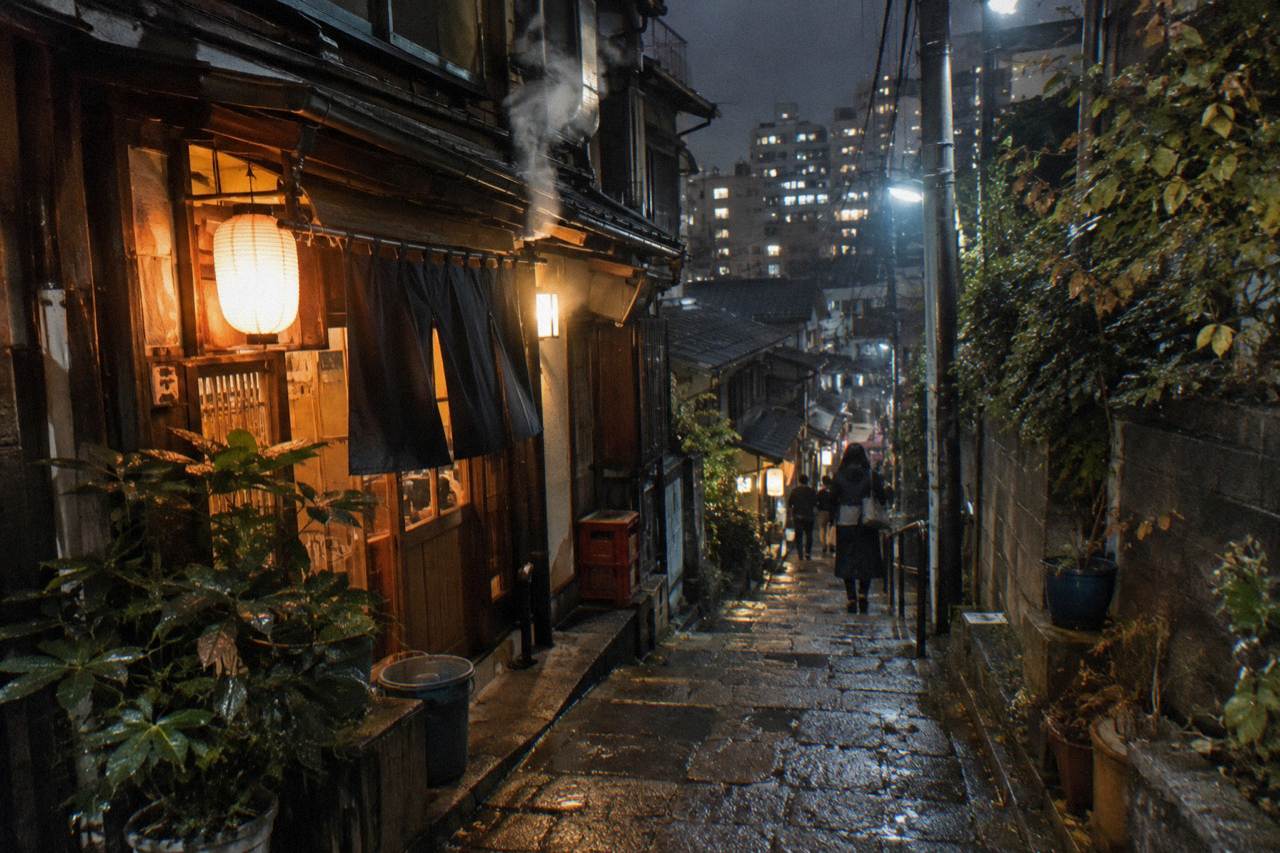 Warm lantern light over a narrow alley of small bar entrances with handwritten signs