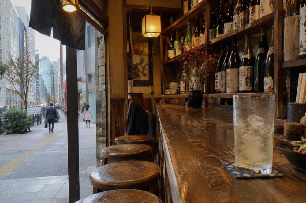 A small izakaya counter with bottles lined on shelves and warm lantern light over wooden stools