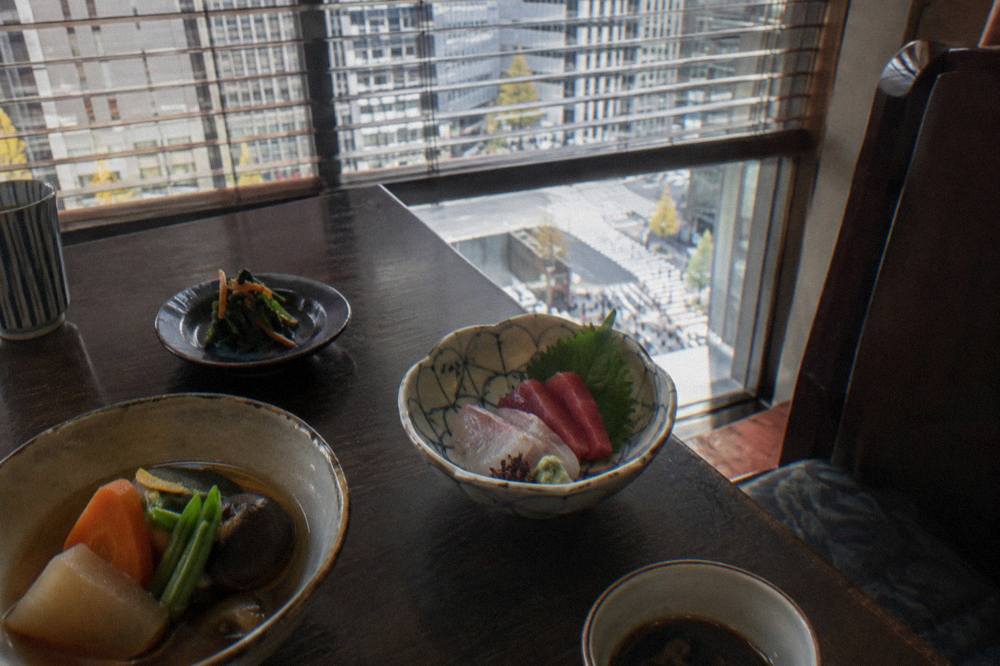 A row of small ceramic dishes holding modest portions of seasonal Japanese food arranged along a dark wooden table