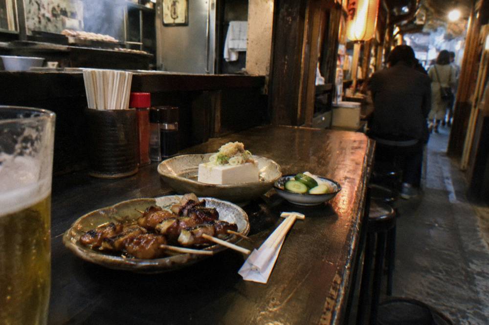 An assortment of small izakaya dishes including skewers, tofu and pickled vegetables spaced along a counter with chopsticks resting beside them
