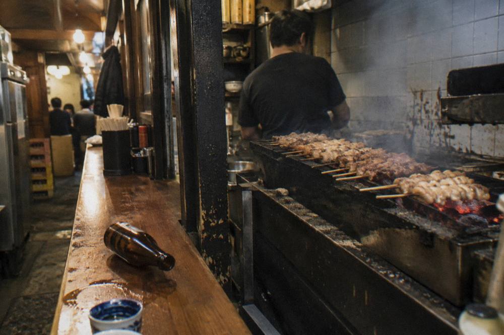 Rows of grilled chicken skewers cooking over charcoal behind a small wooden counter in a narrow lane