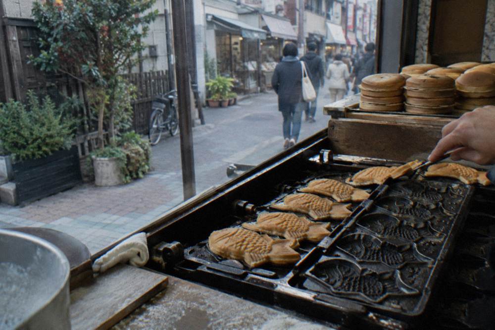 Golden fish-shaped cakes cooling on a hot griddle beside stacks of round pancakes at a small shop window