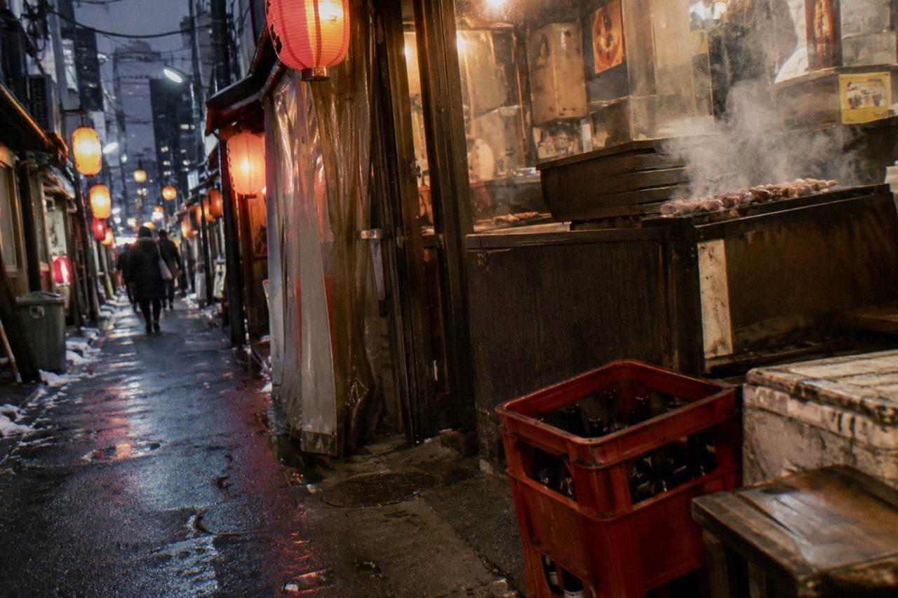 Steam rising from a narrow alley of small grill-front food stalls lit by red lanterns in the evening