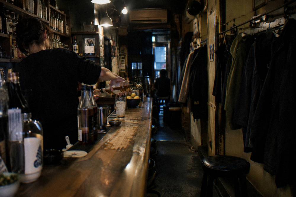 A bartender pouring a drink across a worn wooden counter in a tiny standing bar