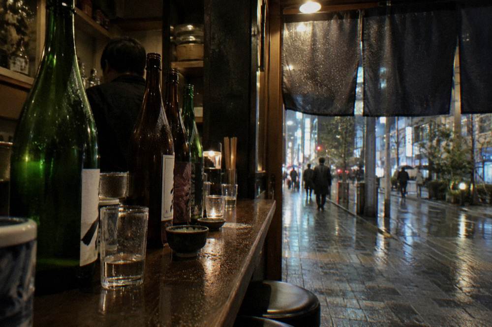 A row of sake bottles and glasses arranged along a dim wooden bar counter