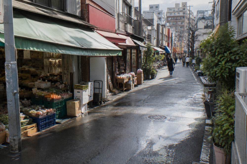 A long shotengai shopping street lined with small grocery and snack shops stretching toward a residential skyline
