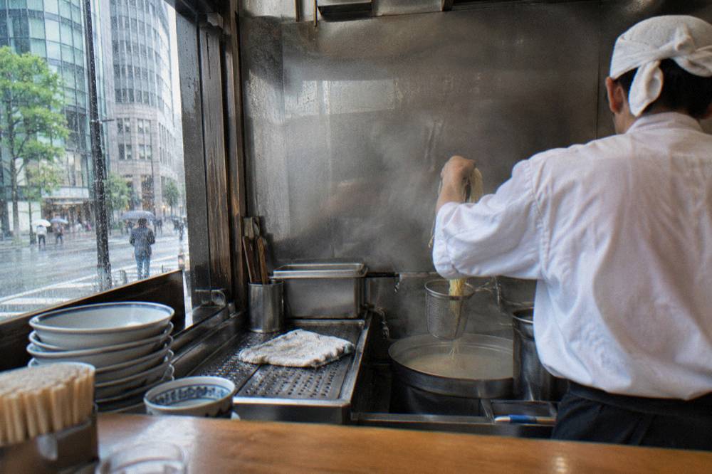 Chef placing noodles into a strainer over steaming broth behind a ramen counter