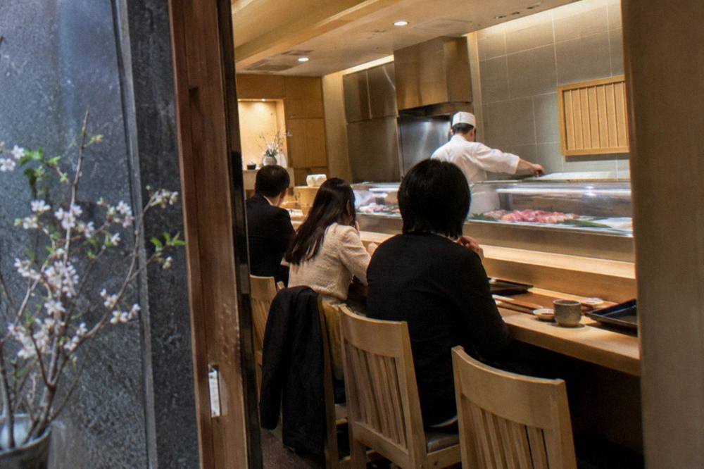 Small group of diners seated at a wooden counter with chef preparing sushi behind the bar