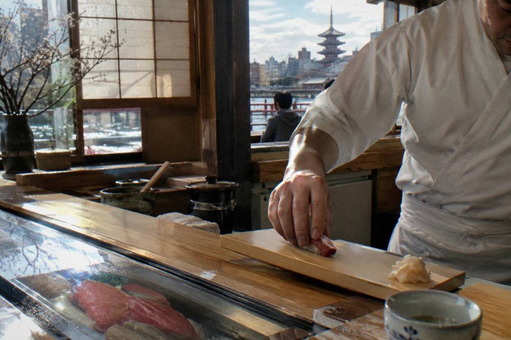 Chef placing a piece of nigiri across a blond wood sushi counter
