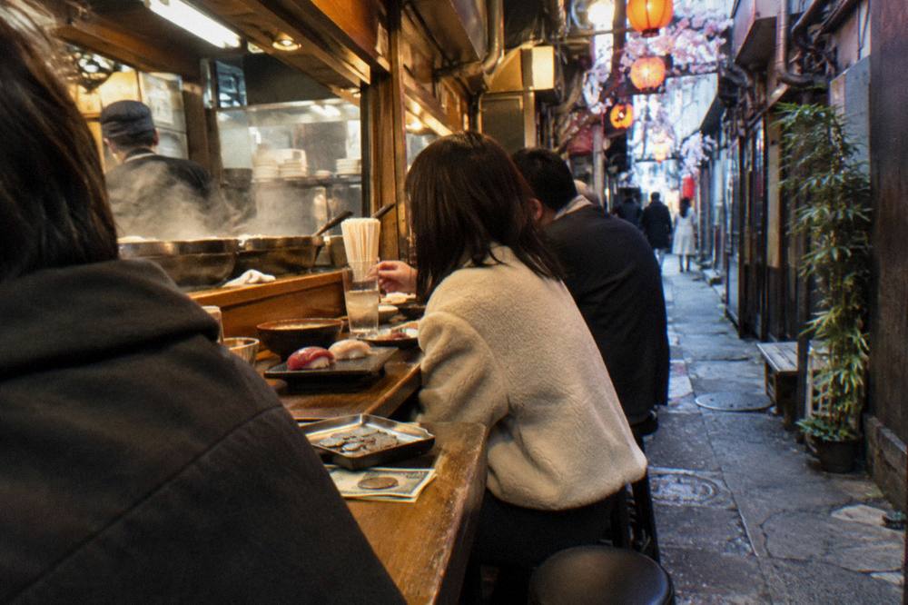 Diners seated at a narrow standing bar counter with sushi plates in front of them