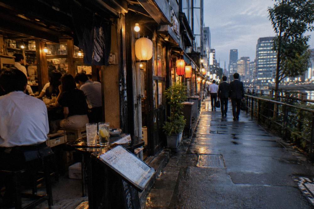 Crowded izakaya alley in the evening with warm light spilling from open doorways onto the pavement
