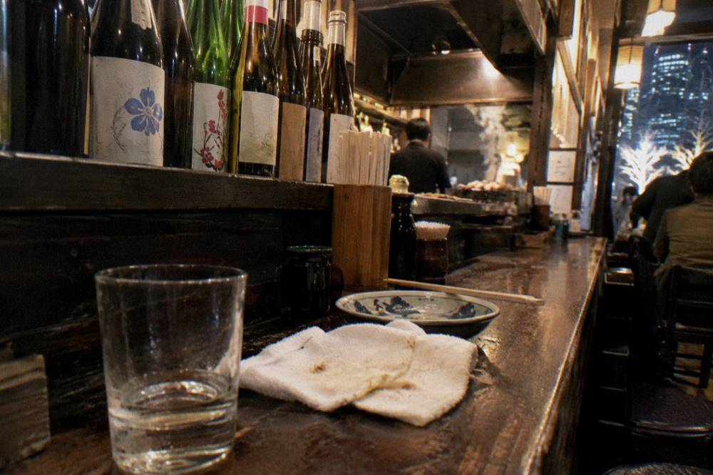 Wooden counter of a small izakaya with sake bottles lined along the back shelf