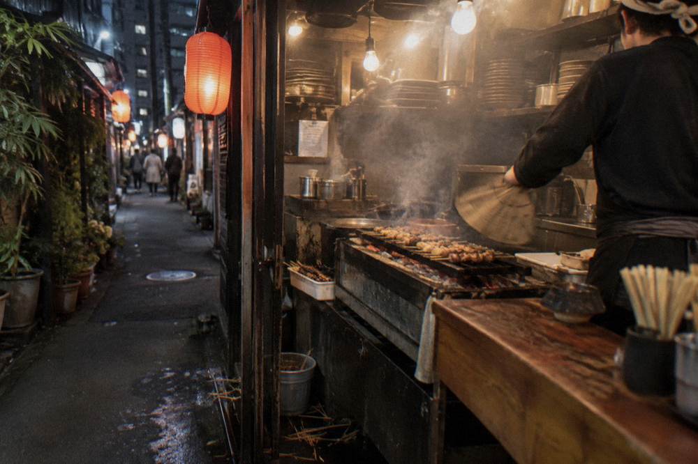 Chef grilling skewers behind a narrow counter in a lantern-lit alley restaurant