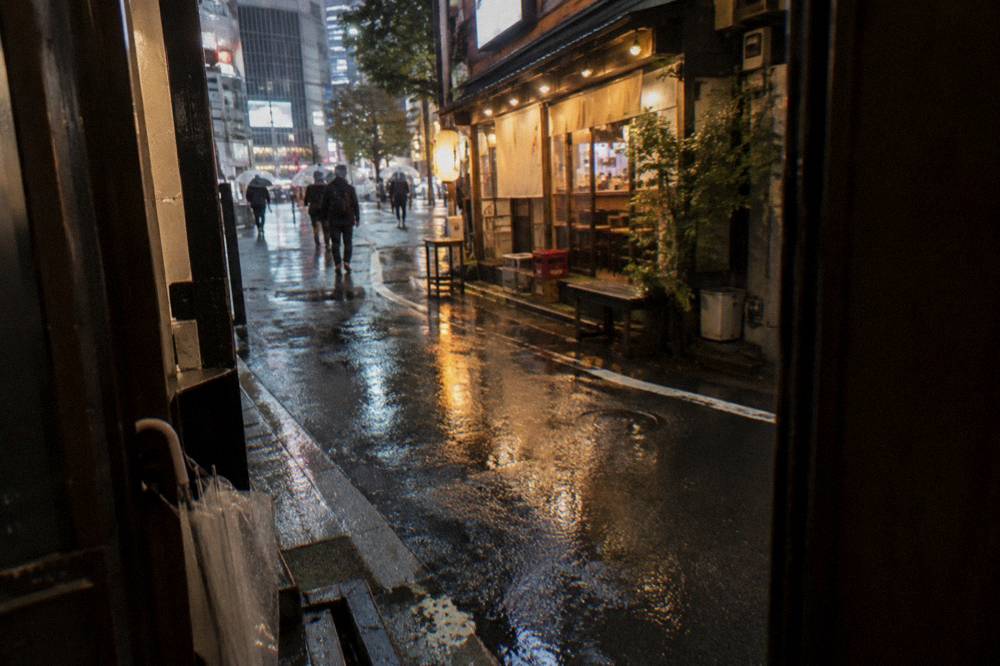 Warm light spilling from open restaurant fronts onto a wet Shibuya side street at night