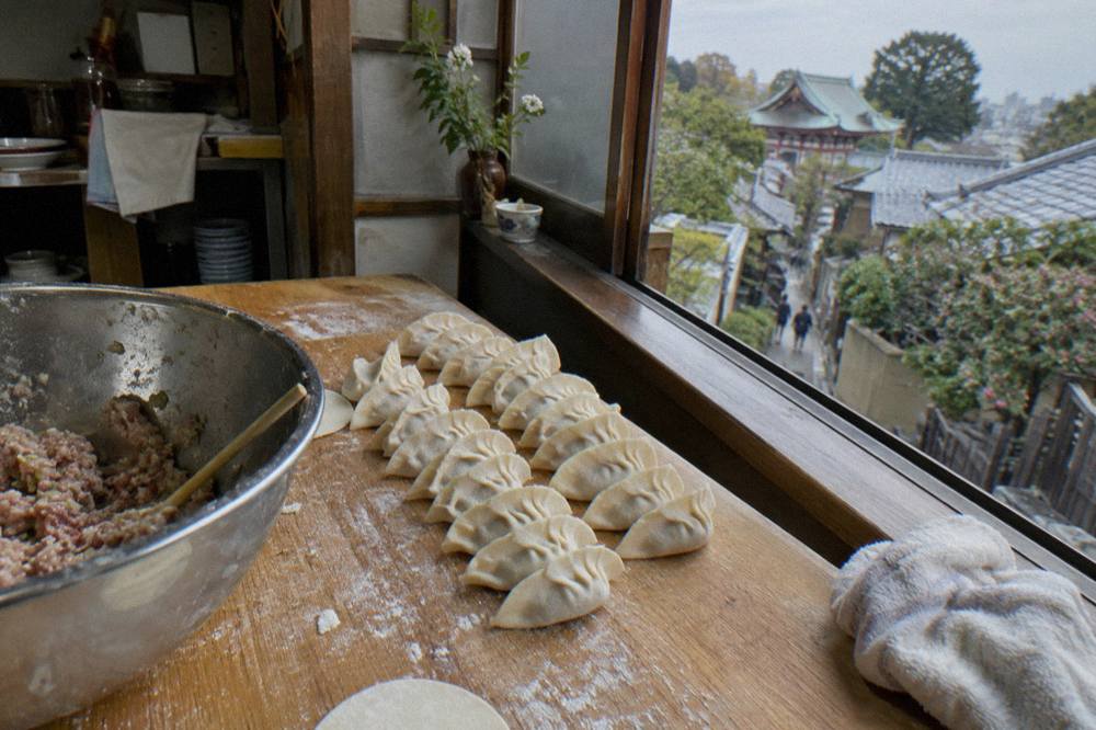 Hands folding gyoza dumplings on a floured wooden kitchen counter beside a bowl of filling.