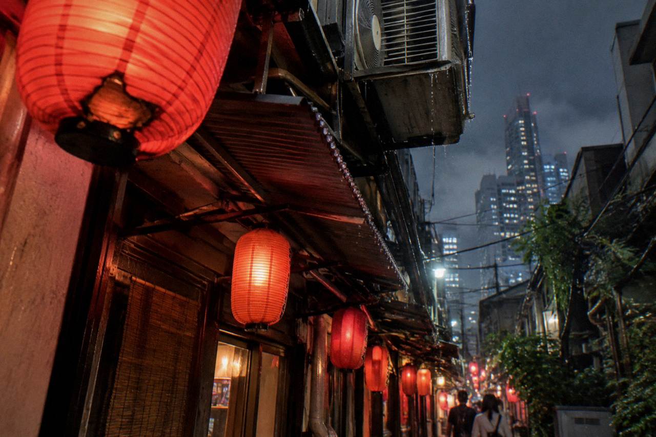 Warm lantern light over a narrow Shinjuku alley of small izakaya counters at night