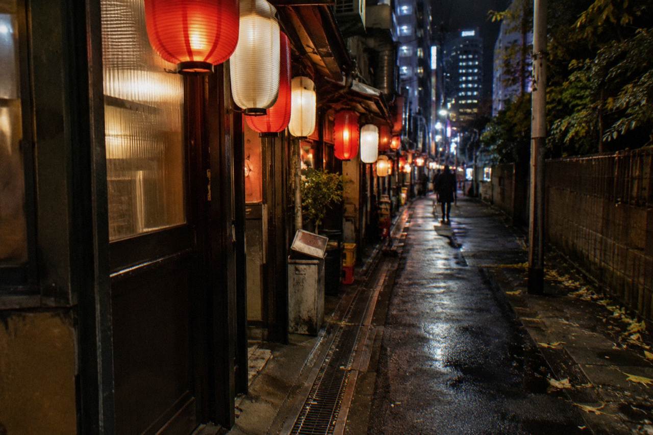 A narrow lantern-lit alley in Tokyo at night with small bar entrances on both sides