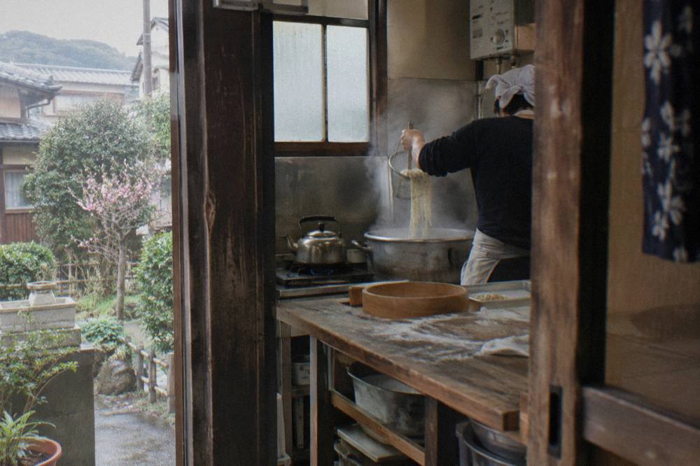 A cook straining fresh ramen noodles over a steaming pot in a small Tokyo kitchen