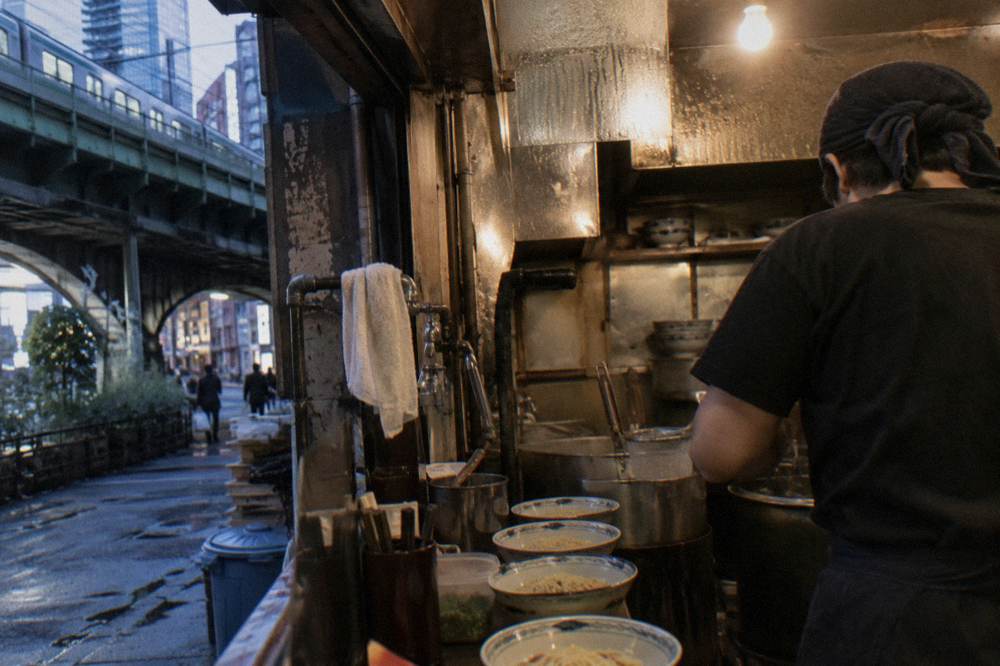 A small restaurant kitchen with a chef portioning noodles into bowls during evening service