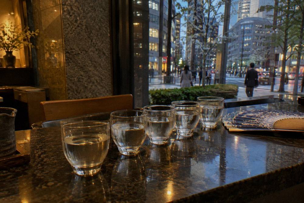 Small tasting glasses of sake arranged in a row on a dark wooden bar top