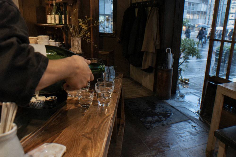 A host pouring clear sake from a bottle into small tasting glasses at a wooden counter