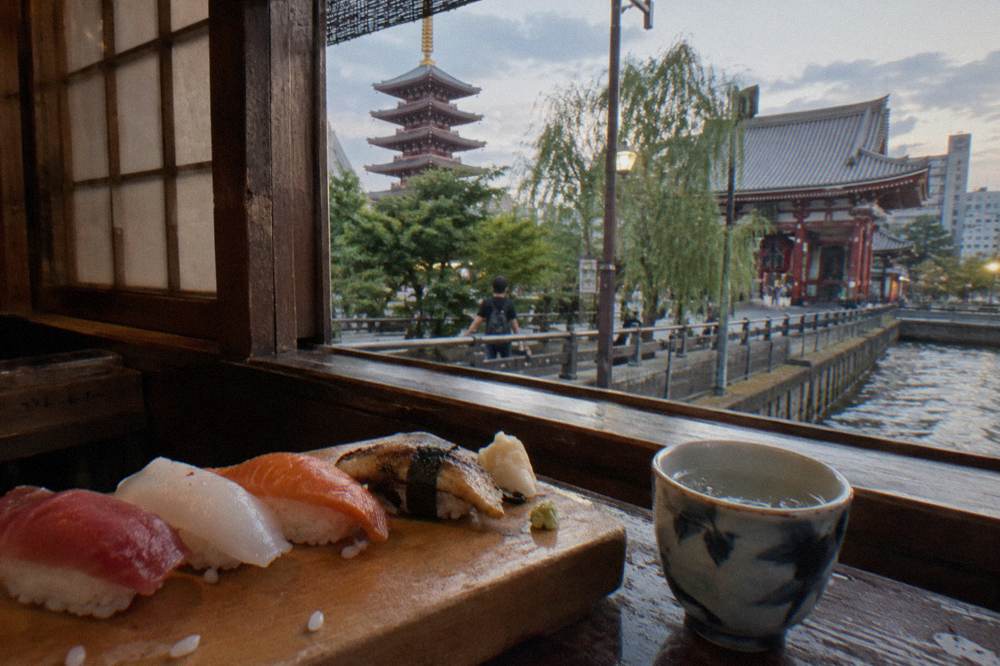 Hand-shaped nigiri sushi pieces resting on a wooden board beside a small cup of sake