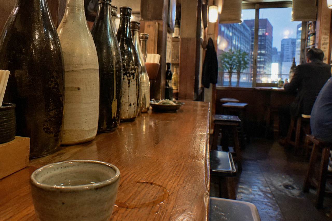 Rows of sake bottles lined up on a wooden bar counter under warm lighting