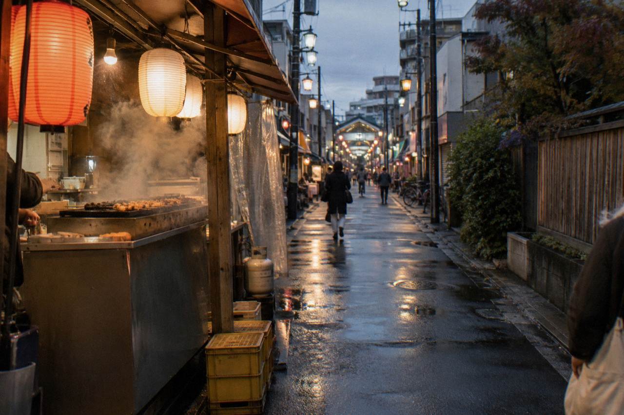 Lantern-lit alley of small food stalls with steam rising from a grill counter in the evening