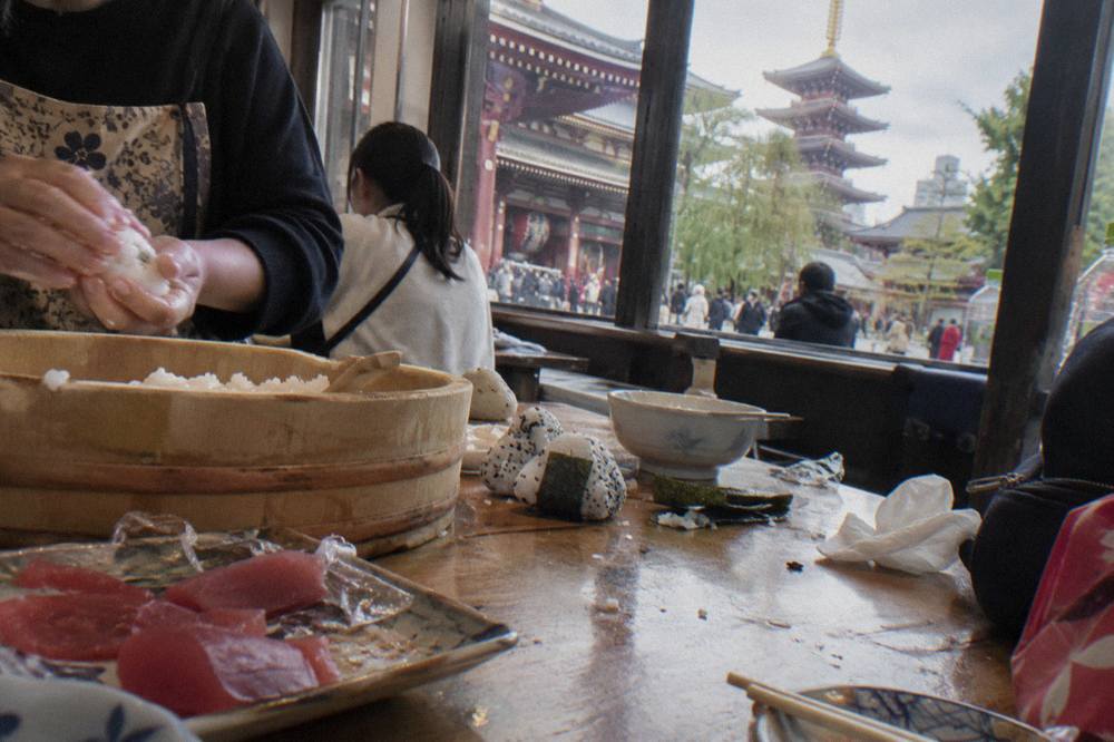 Hands pressing a ball of seasoned rice and a slice of tuna into a nigiri piece on a wooden counter