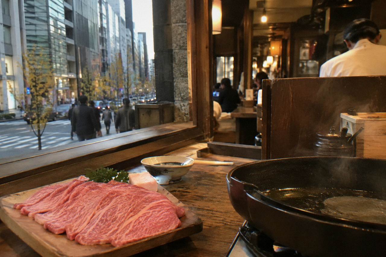Sliced marbled raw beef on a wooden board beside a hot pan in a warm kitchen