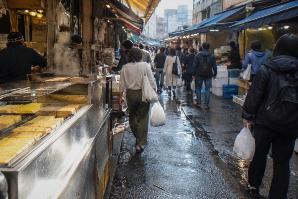 Crowded Tsukiji market lane in late morning with shoppers carrying bags