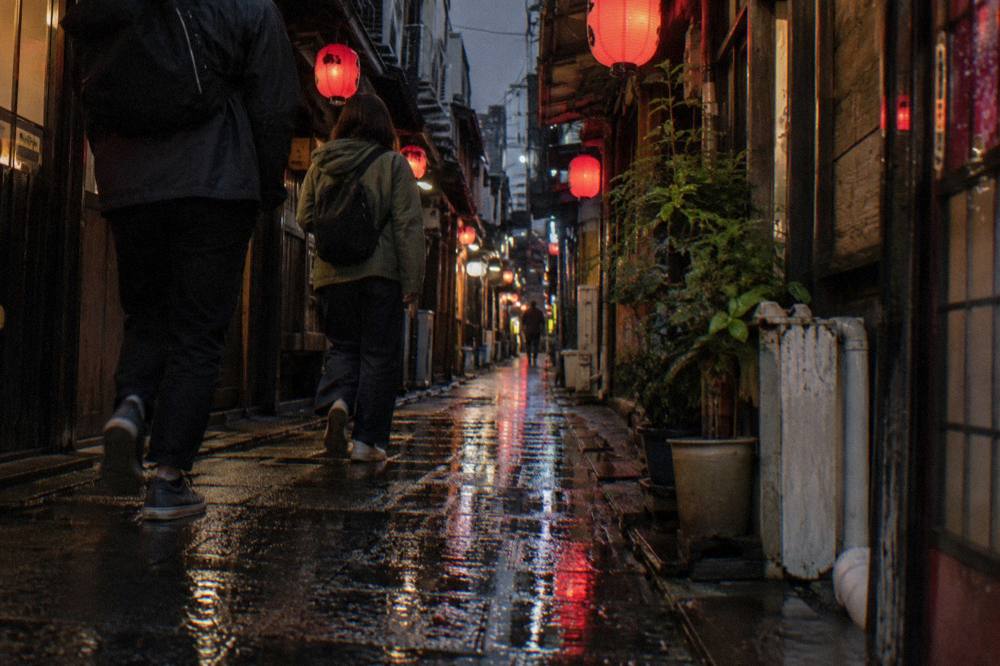 Travellers in casual clothes and walking shoes passing under lanterns in a Tokyo eating district at night