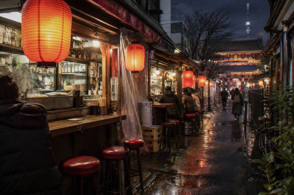 Covered izakaya lane at night with red lanterns glowing over small counter bars