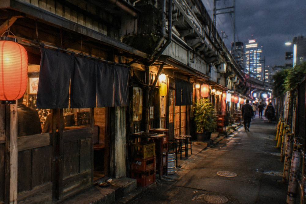 A row of small izakaya storefronts in a Shibuya side street at night with warm lantern light