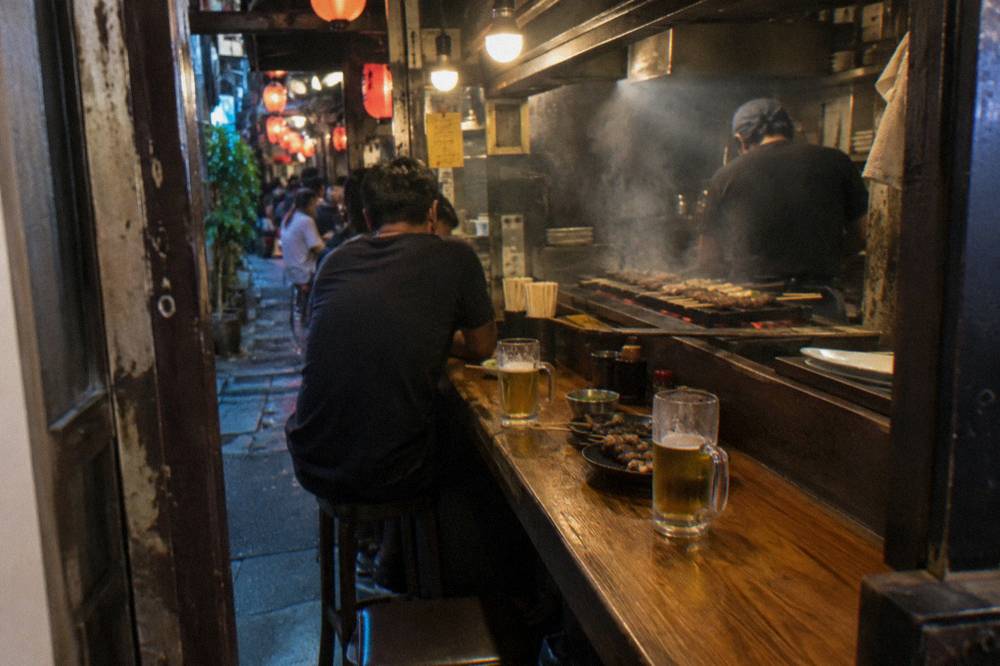 A small izakaya counter with grilled skewers and glasses of beer set along wooden boards