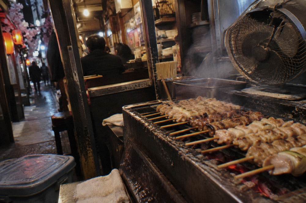 Charcoal grill loaded with chicken skewers inside a cramped counter restaurant