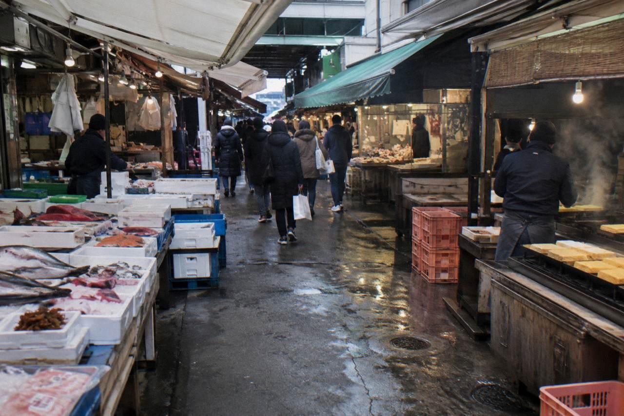 Wide morning view down the covered arcade of Tsukiji Outer Market in Tokyo, stalls of fresh fish and tamagoyaki under white canopies